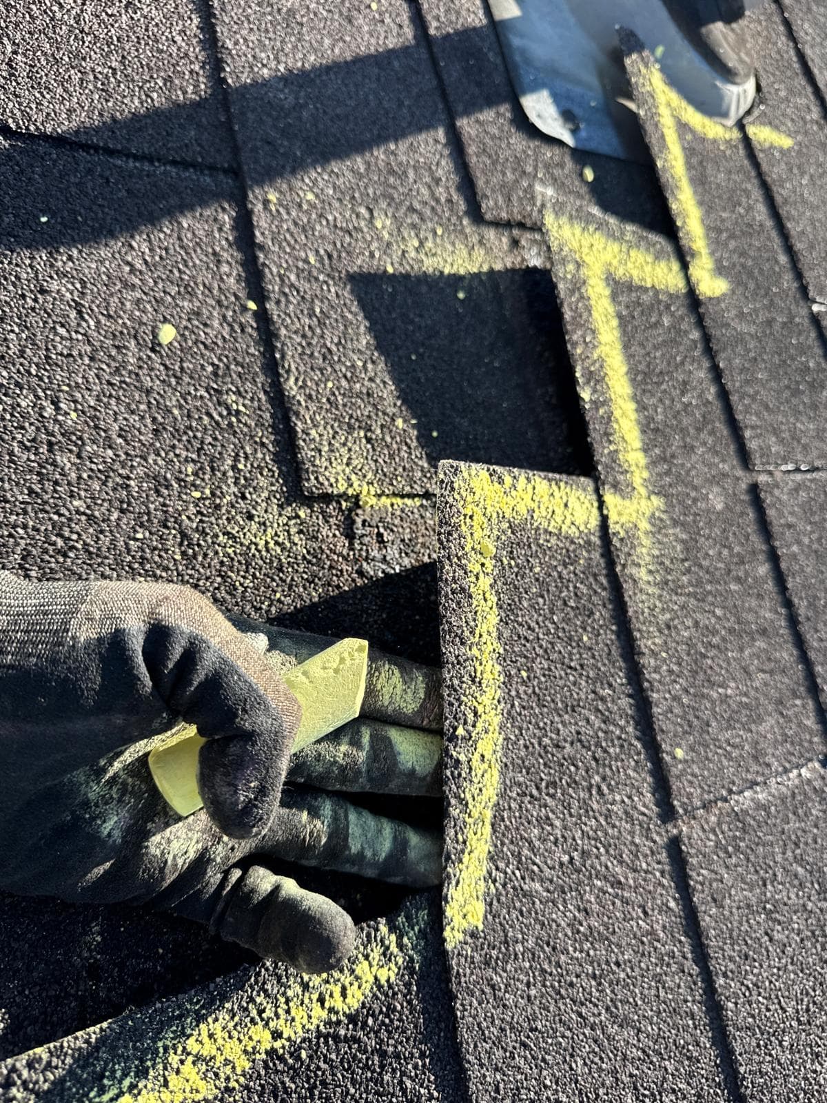 Gloved hand lifting a dark asphalt shingle marked with yellow chalk during a roof inspection.